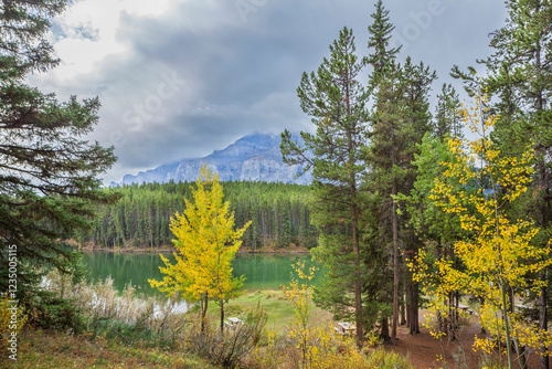 A scenic view of a serene lake surrounded by evergreen forests and golden autumn trees, with majestic mountains in the background under a cloudy sky in Canada.