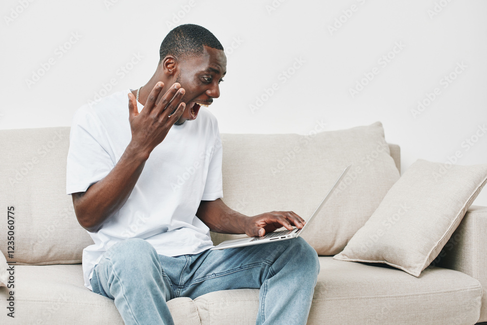Excited young Black man showing enthusiasm while using a laptop on a cozy couch, embodying the joy of online communication and connection Excited young Black man showing enthusiasm while using a laptop on a cozy couch, embodying the joy of online communication and connection