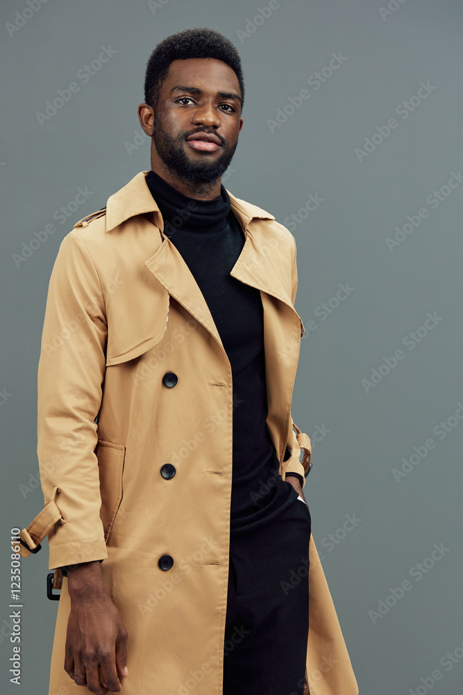 Fototapeta premium Stylish African man in beige trench coat exuding confidence against a gray background, projecting a modern fashion vibe perfect for seasonal trends