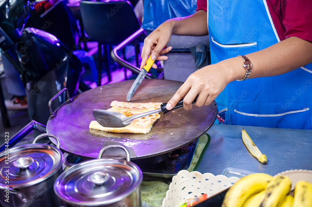 Fototapeta premium Street vendor preparing traditional Thai banana roti on a hot griddle at a bustling night market, showcasing authentic flavors and culinary skills