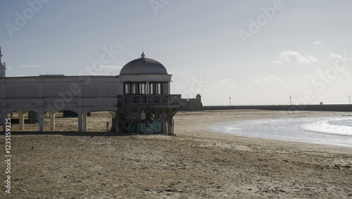 A historic building with a domed tower overlooks a sandy beach with gentle waves. The architecture features arched walkways and a classic design, with a clear blue sky above.

 photo