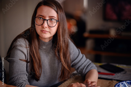 A woman with glasses, wearing a gray sweater, concentrating on a creative project indoors. The scene captures focus and dedication. photo