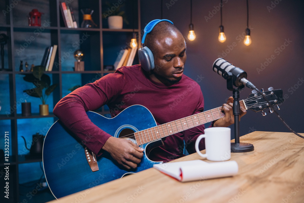 Obraz premium African American male musician wearing headphones, playing acoustic guitar while recording podcast, seated at desk in home studio setting