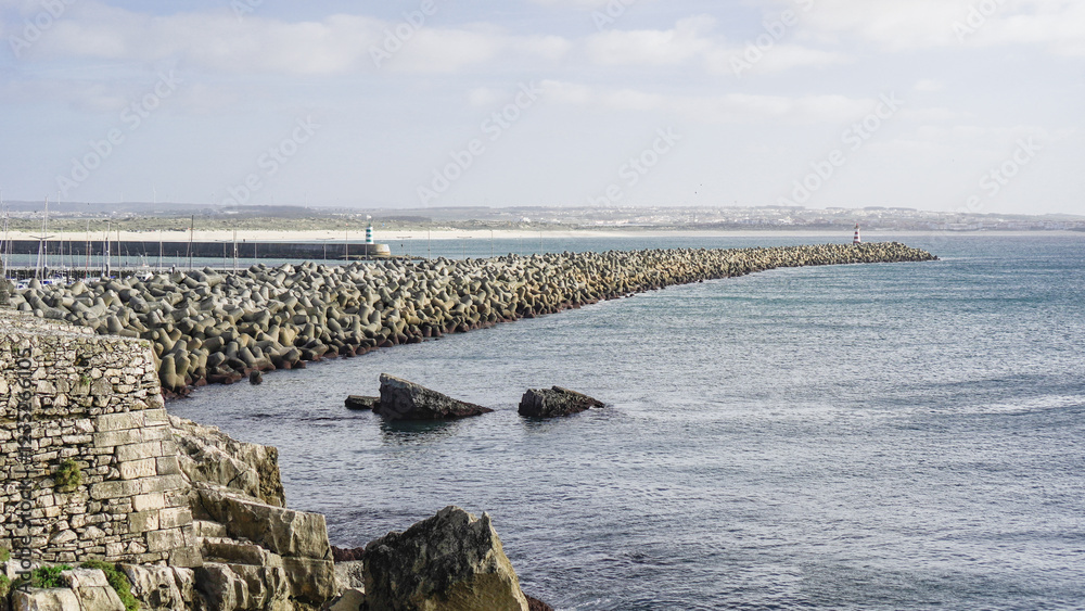 A long stone breakwater extends into the calm blue sea, protecting the shore. Large rocks emerge from the water, while the distant coastline stretches under a clear sky.
A long stone breakwater extends into the calm blue sea, protecting the shore. Large rocks emerge from the water, while the distant coastline stretches under a clear sky.