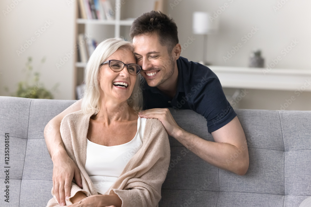 Fototapeta premium Visiting mom. Affectionate young adult son hug smiling elder aged mother sitting on couch with warmth care lean head to her with closed eyes sincere smile feel gratitude to single parent grown him up