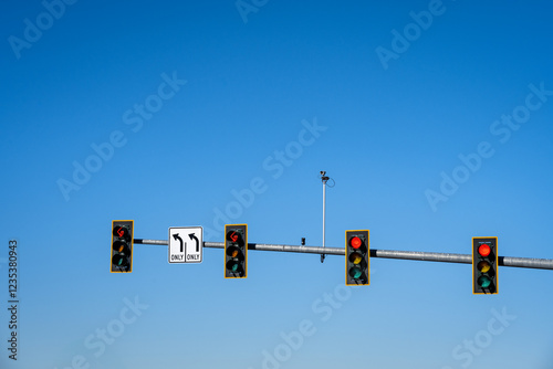 Overhead traffic light for four lanes, two left turn only lanes, sign and red arrow light, and two lanes with red stop light, traffic camera, sunny winter blue sky day
