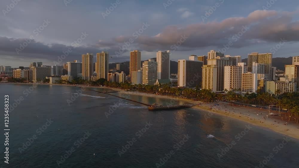 Aerial view of Waikiki Beach in Honolulu, Hawaii, during sunset. High rise skyline, ocean waves, and lush mountains create a serene coastal scene. Aerial view of Waikiki Beach in Honolulu, Hawaii, during sunset. High rise skyline, ocean waves, and lush mountains create a serene coastal scene.