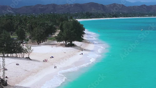 Aerial view of a beach on Oahu island, Hawaii, with turquoise waters, white sand, palm trees, mountain ranges, and kite surfers gliding over the waves.