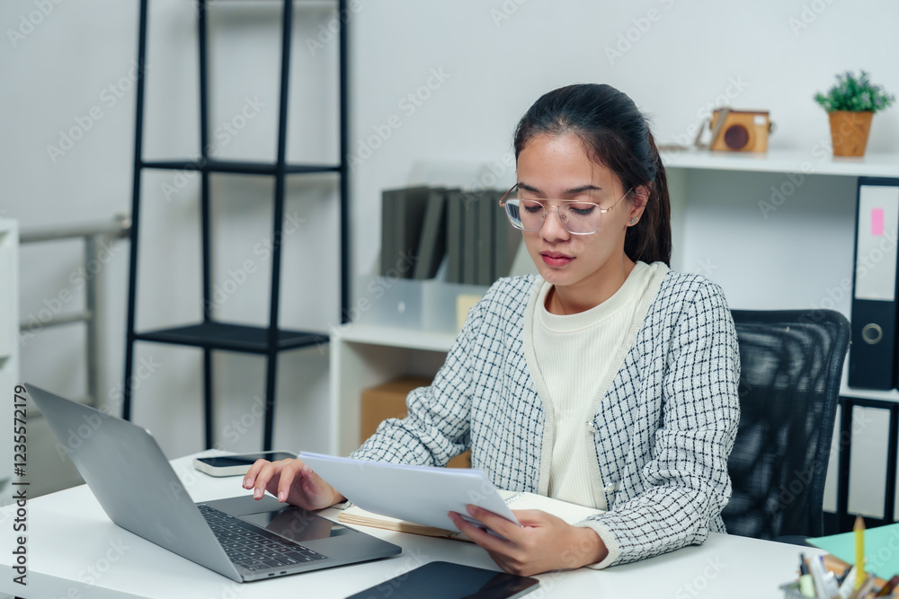 Asian female office worker wearing glasses reviewing documents at a modern desk. Professional workspace with laptop, office supplies, and organized shelves creates a productive corporate environment. Asian female office worker wearing glasses reviewing documents at a modern desk. Professional workspace with laptop, office supplies, and organized shelves creates a productive corporate environment.