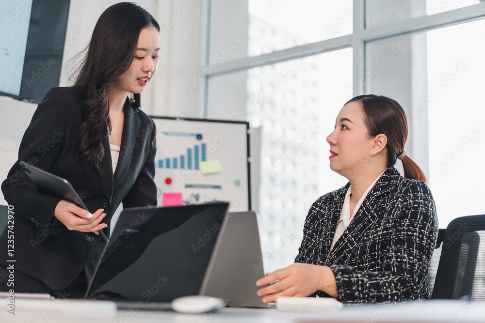 Two businesswomen discussing project in modern office with laptop and charts in background Two businesswomen discussing project in modern office with laptop and charts in background