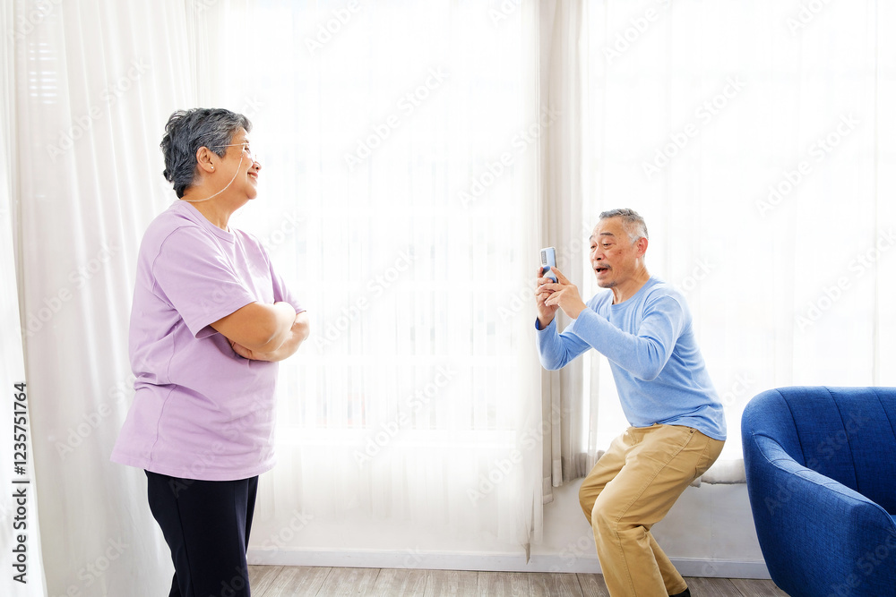Fototapeta premium A senior couple shares a joyful moment at home as the husband playfully takes a photo of his wife with a smartphone. Bright room, happy expressions highlight love, companionship, and fun in retirement
