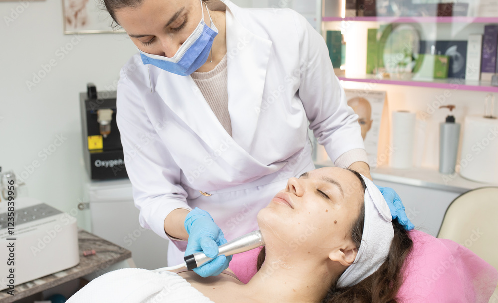 Naklejka premium Woman is getting her face done by a doctor. The doctor is wearing a white coat and a blue mask. The woman is lying on a bed and the doctor is using a device to clean her face