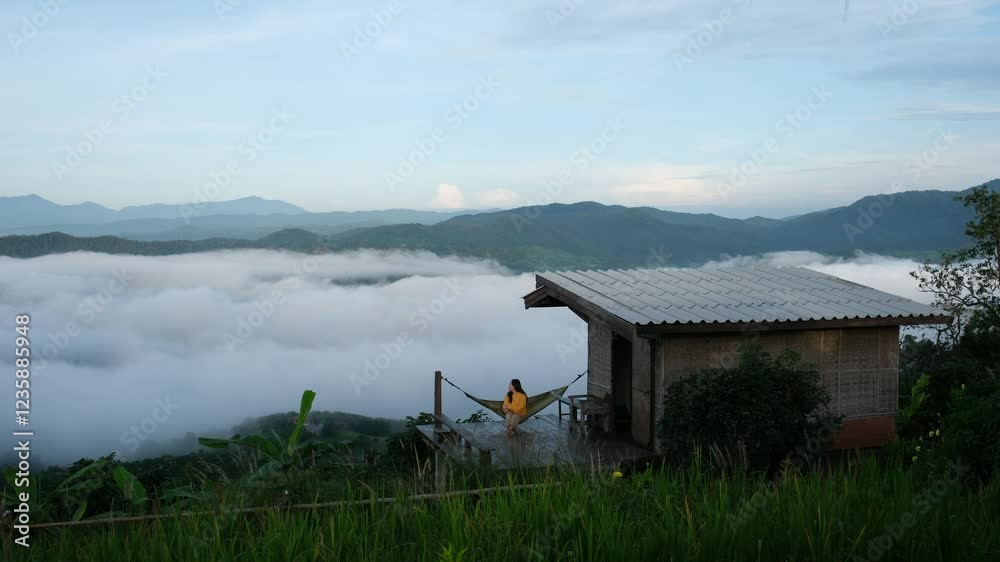 A woman sitting on hammock on wooden house terrace while looking at a nature view and sea of fog A woman sitting on hammock on wooden house terrace while looking at a nature view and sea of fog