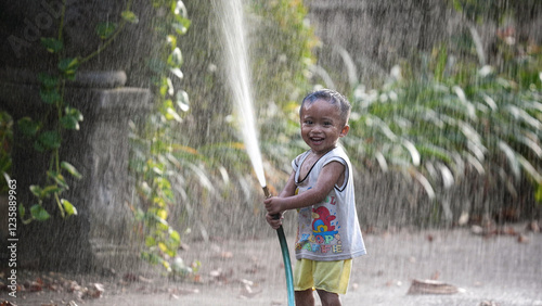 Happy little boy play with water hose. Smile asian child have fun. Cheerful childhood. Laughing kid enjoy rain splash. Joyful wet toddler. Cute baby relax outdoor slow motion. Nice hot day. Cool rest. photo