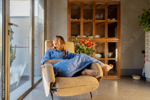 A woman in a blue linen outfit relaxes in a vintage armchair near a sunlit window. The modern interior, wooden cabinet, and fresh flowers create a warm, cozy, and inspiring atmosphere of slow living photo