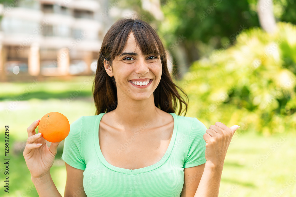Young woman holding an orange at outdoors pointing to the side to present a product Young woman holding an orange at outdoors pointing to the side to present a product