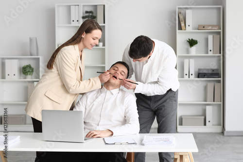 Young people drawing mustache on their sleeping colleague's face in office. April Fools' Day celebration