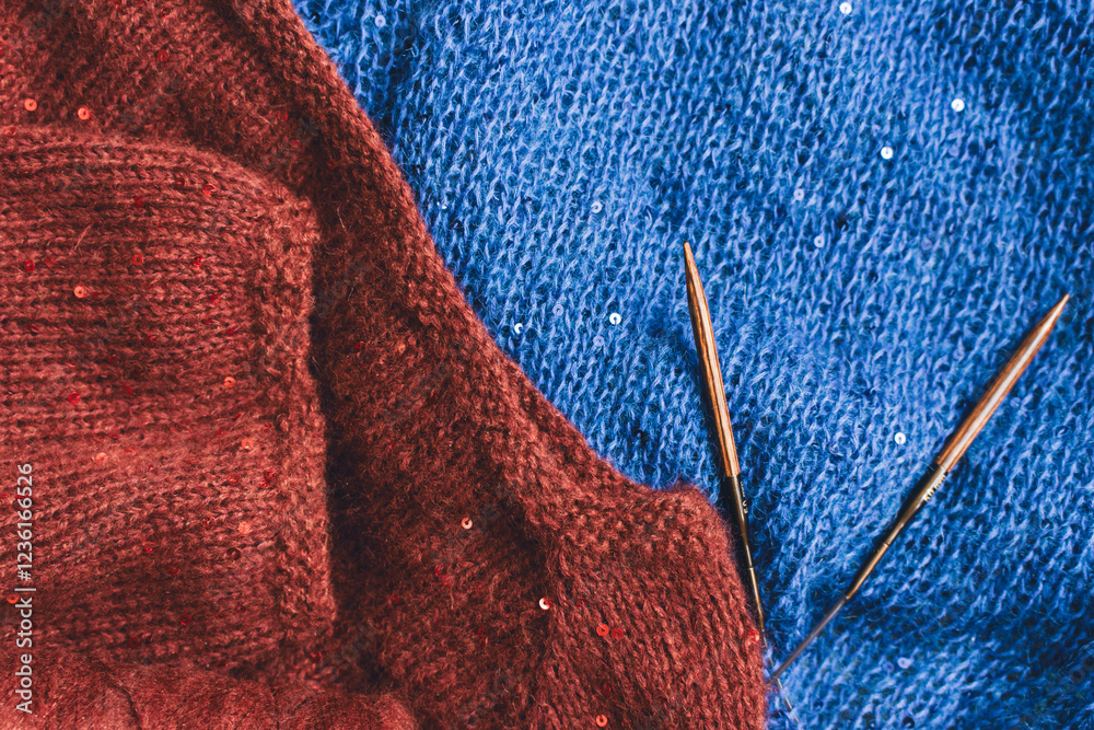Red and blue knitted sweater with sequins and knitting needles. Sequinned knitwear on wooden needles. Sequin pattern in needlework. Knitting hobby. Knitter work. Sequins texture in knitwear.
