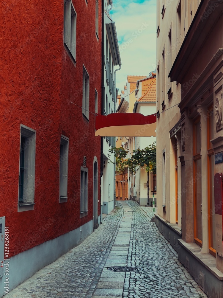 Fototapeta premium A charming narrow alley with cobblestone pavement, framed by colorful historic buildings. The red facade contrasts beautifully with the muted tones, creating a picturesque European street scene.
