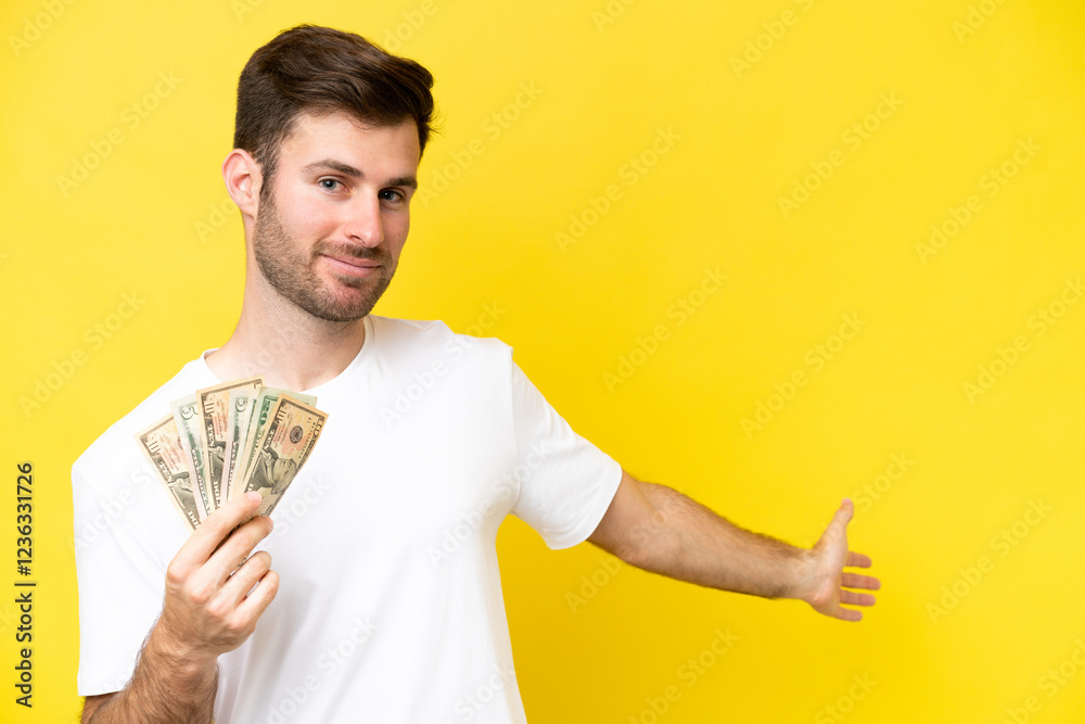 Young caucasian man taking a lot of money isolated on yellow background extending hands to the side for inviting to come Young caucasian man taking a lot of money isolated on yellow background extending hands to the side for inviting to come