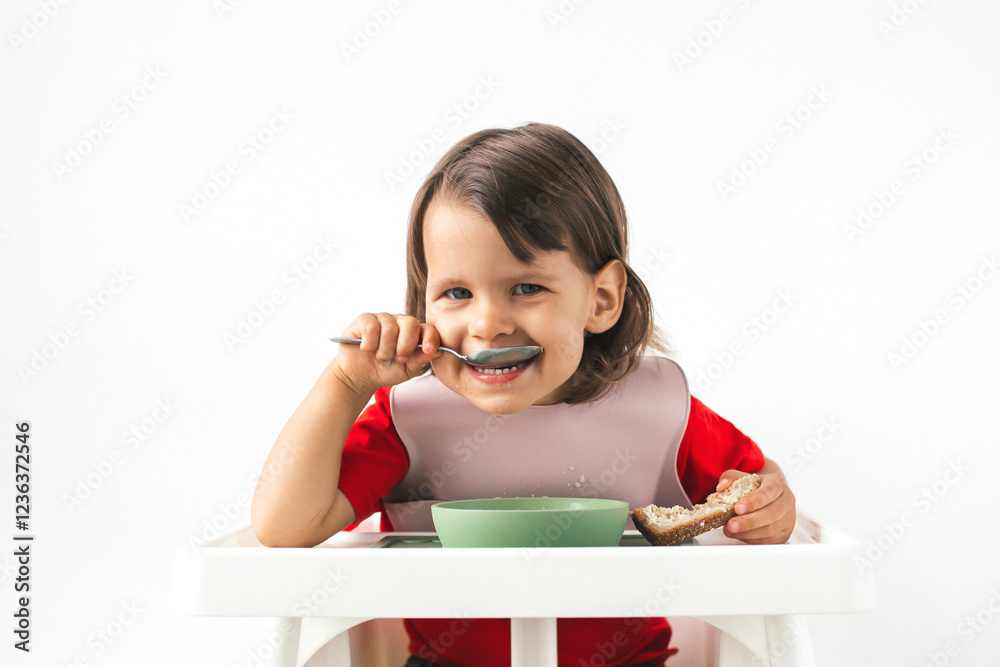 Happy funny toddler in a red shirt and pink bib sitting in a high chair, eating with a spoon and holding bread. The child s joyful smile and bright eyes create a warm and cheerful atmosphere Happy funny toddler in a red shirt and pink bib sitting in a high chair, eating with a spoon and holding bread. The child s joyful smile and bright eyes create a warm and cheerful atmosphere