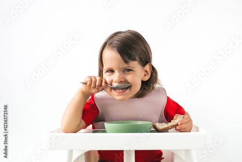 Happy funny toddler in a red shirt and pink bib sitting in a high chair, eating with a spoon and holding bread. The child s joyful smile and bright eyes create a warm and cheerful atmosphere photo