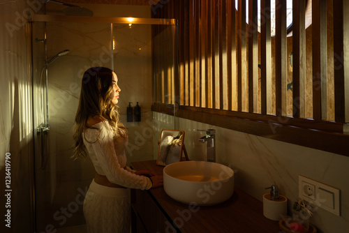 Woman basking in golden sunlight streaming through wooden slats, illuminating minimalist bathroom with tranquil, soft radiance