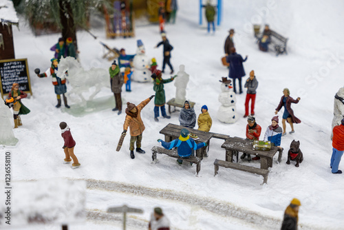 Festive winter scene with miniature people enjoying outdoor activities, building snowmen, sitting at wooden tables, observing ice sculptures, and playing in the snow