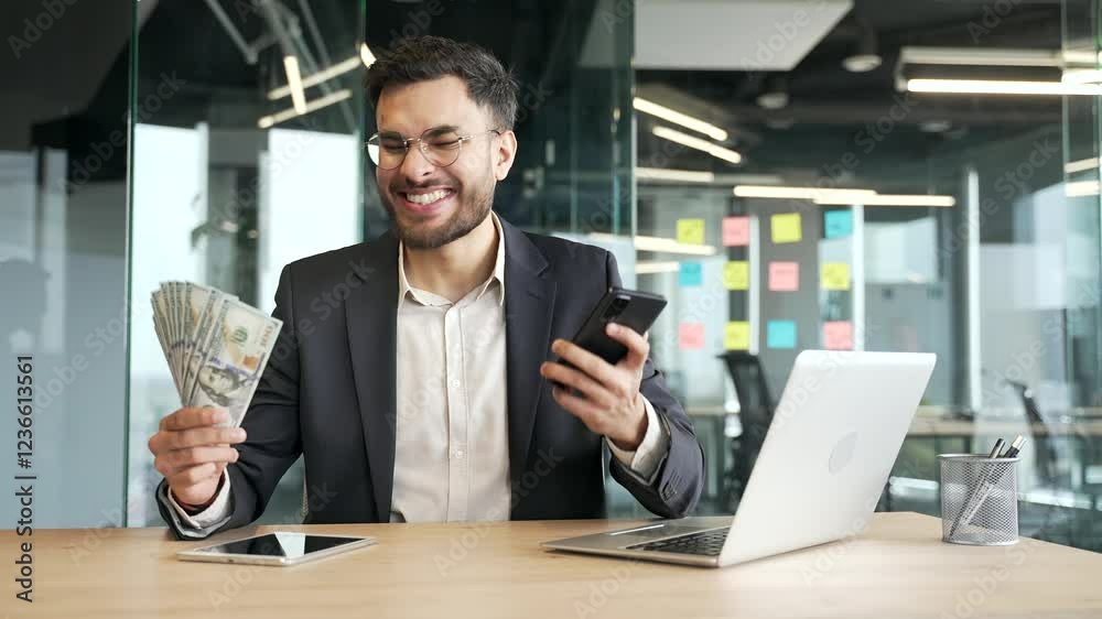 Happy businessman in a formal suit holding a fan of cash money dollar bills and phone in business office looking at camera. Male is satisfied with earning money using an application on his smartphone Happy businessman in a formal suit holding a fan of cash money dollar bills and phone in business office looking at camera. Male is satisfied with earning money using an application on his smartphone