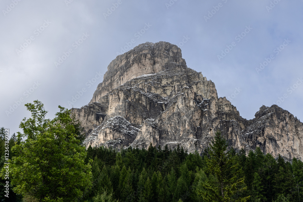 Sassongher mountain, with its steep, rocky cliffs, towers above a dense forest of evergreens, creating a stunning contrast against the clear sky and scattered snow Sassongher mountain, with its steep, rocky cliffs, towers above a dense forest of evergreens, creating a stunning contrast against the clear sky and scattered snow