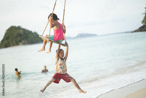 Two children have a joyful moment swinging on a beach, surrounded by scenic views of the ocean and nature, reflecting happiness and carefree childhood memories. photo