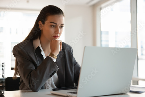 Focused pensive young businesswoman working at laptop in office, touching chin, thinking on online project, Internet marketing statistics, sitting at workplace table with desktop, using technology