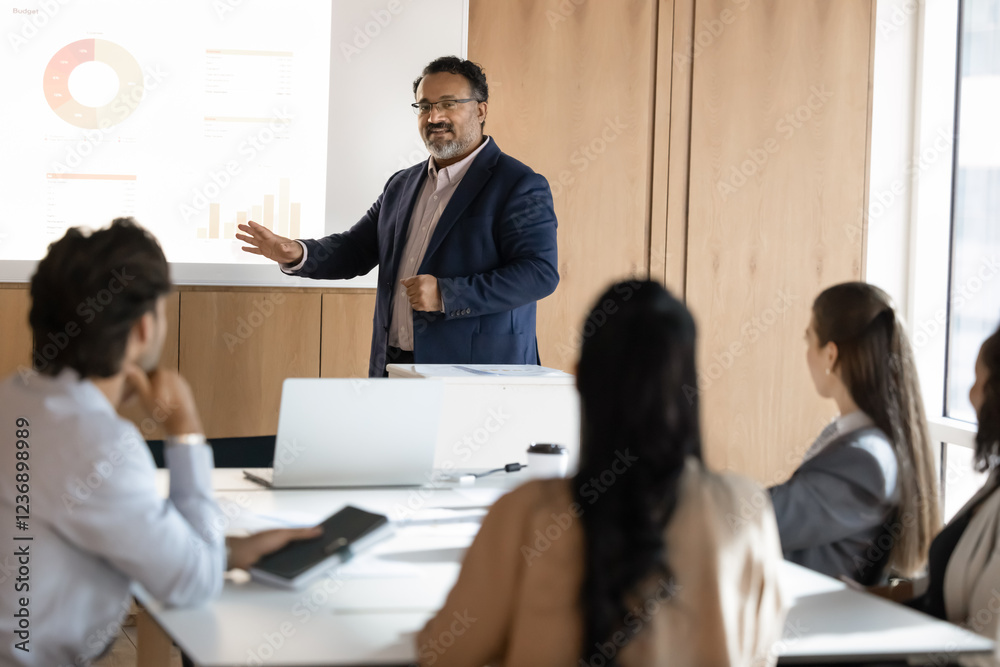 Older Indian diverse team leader man presenting marketing data, financial sales report on whiteboard to colleagues, speaking at screen, holding presentation meeting with coworkers Older Indian diverse team leader man presenting marketing data, financial sales report on whiteboard to colleagues, speaking at screen, holding presentation meeting with coworkers