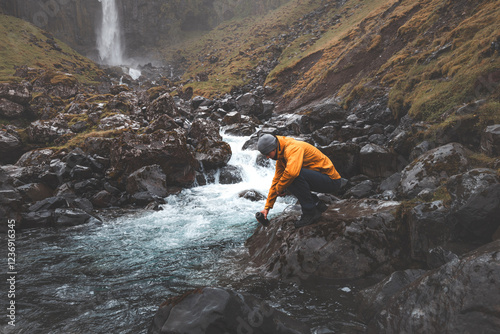 Traveler in an orange jacket kneels by a rushing stream near Grundarfoss waterfall on the Snaefellsnes Peninsula, Iceland, filling a bottle with fresh, crystal-clear glacial water photo