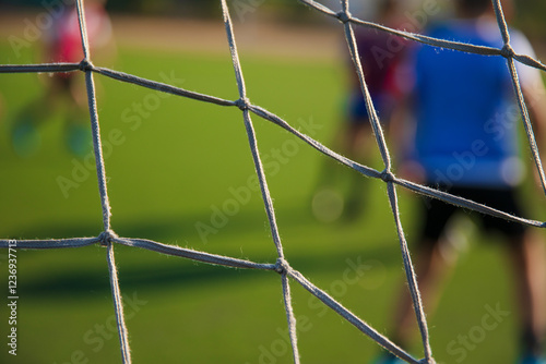 The photo shows a close-up of a soccer net with blurred players in the background, engaged in a game on a green turf field. The focus is on the net, with the players' movements softly visible. photo