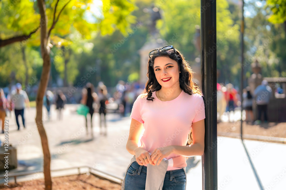 Fototapeta premium Cheerful young Latina woman with wavy hair, pink t-shirt, and jeans leans on a lamppost in Alameda Central, Mexico City, enjoying a sunny day surrounded by nature and people.