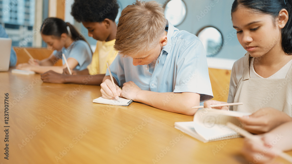 Young smart teenager writing and talking about topic at classroom. Diverse highschool student taking a note in other notebook while sitting with multicultural friends. Creative education. Edification. Young smart teenager writing and talking about topic at classroom. Diverse highschool student taking a note in other notebook while sitting with multicultural friends. Creative education. Edification.