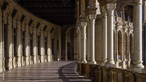 A beautiful arched corridor with elegant columns casts long shadows on the ground. The historic architecture, warm tones, and intricate details create a timeless atmosphere.

