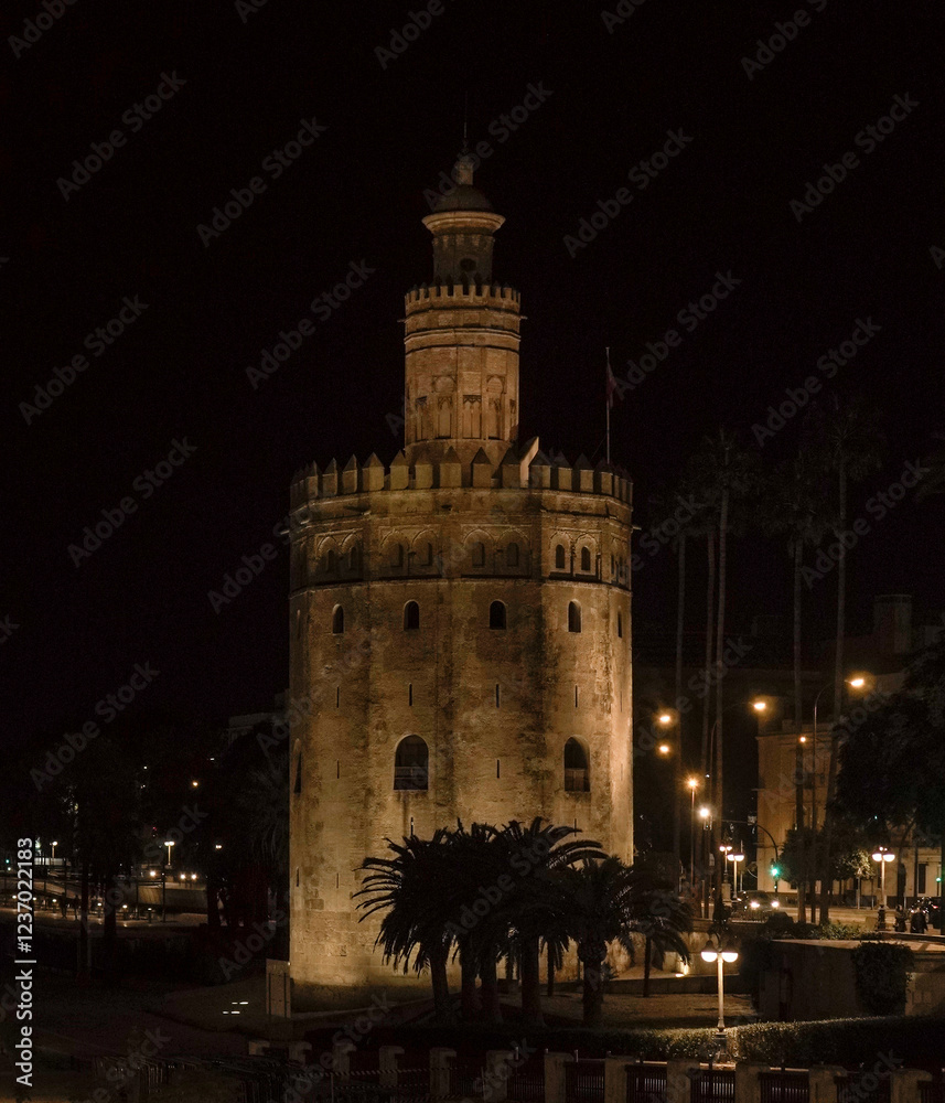 Obraz premium A historic tower stands illuminated at night, overlooking a charming street with outdoor cafés and pedestrians. Warm streetlights add to the cozy, atmospheric cityscape.