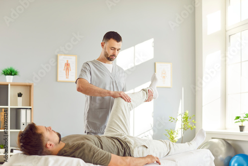 Physician or physiotherapist providing physical therapy and healing treatment for leg recovery to young man on couch. Professional therapist helping male patient with rehabilitation after knee injury.