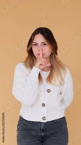 Formal Caucasian young adult woman showing hush gesture with finger on lips on beige background. Concept of a secret. Vertical video.