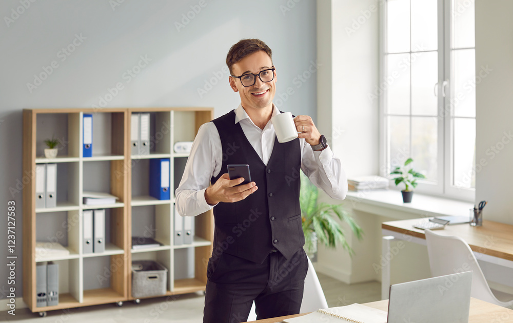Happy, cheerful, friendly young business man in eyeglasses standing in modern office, starting his day with fresh coffee drink, holding mobile phone, looking at camera and smiling Happy, cheerful, friendly young business man in eyeglasses standing in modern office, starting his day with fresh coffee drink, holding mobile phone, looking at camera and smiling