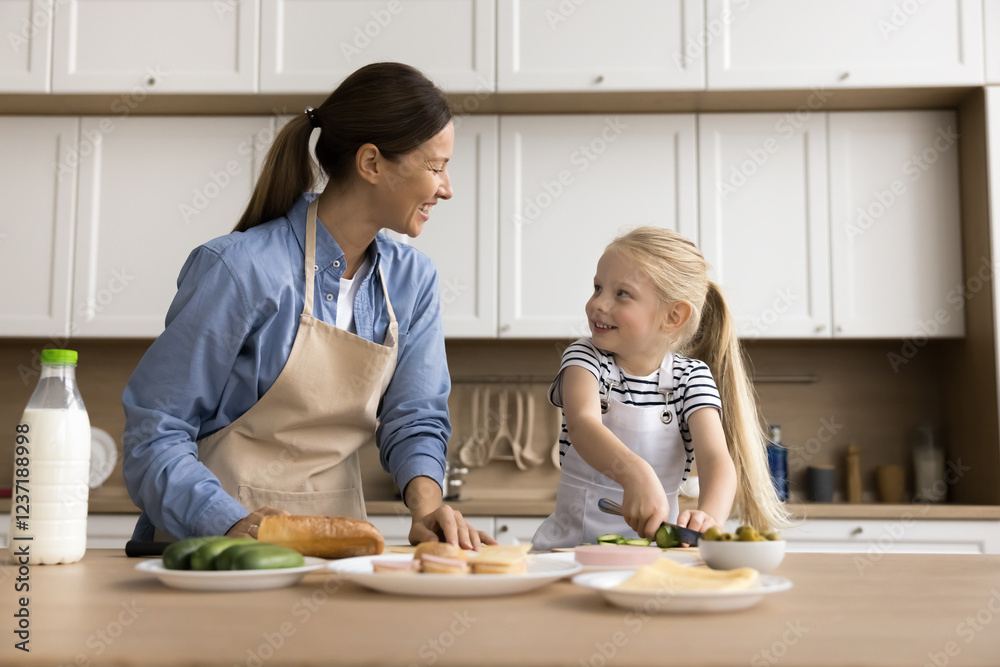 Lovely kid enjoy cookery time with loving mother, wear aprons standing at dining table countertop cutting cucumbers for sandwiches, talking, smiling spend quality time together in modern cozy kitchen Lovely kid enjoy cookery time with loving mother, wear aprons standing at dining table countertop cutting cucumbers for sandwiches, talking, smiling spend quality time together in modern cozy kitchen