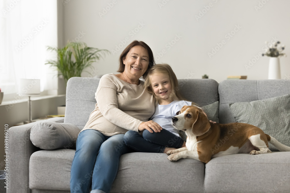 Lovely kid sits on sofa with older grandmother and cute beagle puppy, smile, looking at camera, exuding warmth and togetherness. Multigenerational family ties, understanding and attachment, portrait
