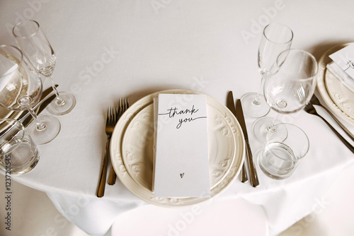 Elegant wedding table setting with a thank you note placed on a decorative white plate, surrounded by fine glassware and silver cutlery.