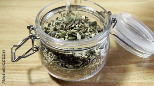 Closeup slow motion shot of mixed edible seeds (sunflower, pumpkin, hemp and linseed) pouring into an open glass storage jar, with an airtight lid and metal clasp. 