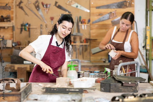 Girl is busy in carpentry workshop. Process wooden billet, sand it, applies pattern with cutter. Related stages of working with devices tools. In workshop girl stand with work gloves in hands photo