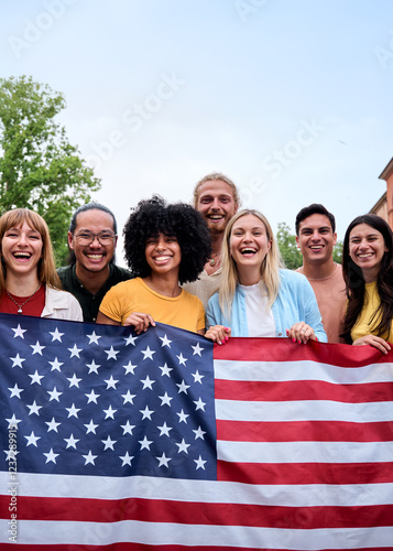 Vertical. Multiracial group of friends holding the American flag with pride, celebrating Independence Day outdoors. Joyful atmosphere with a sense of unity and patriotism in a vibrant summer setting