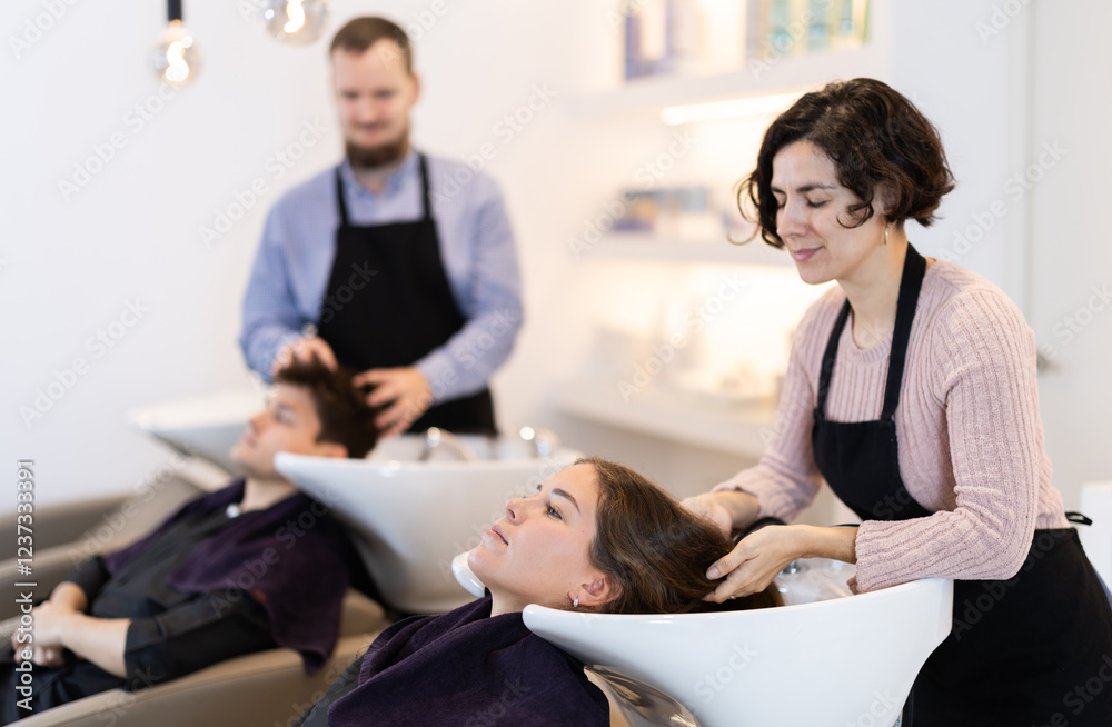 Obraz premium Young woman stylist washes female clients hair, prepares it for cutting and styling. Barbershop visitor placed head in sink, enjoying process of hair care