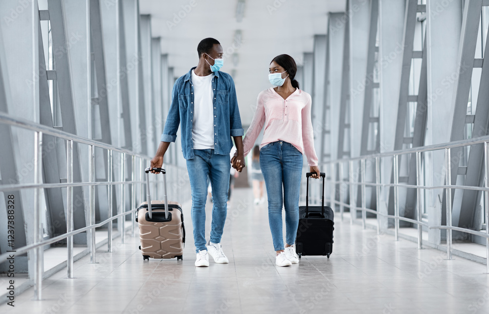 Obraz premium Coronovirus Travels. Black couple in masks walking with luggage at airport building, holding hands and looking at each other, copy space
